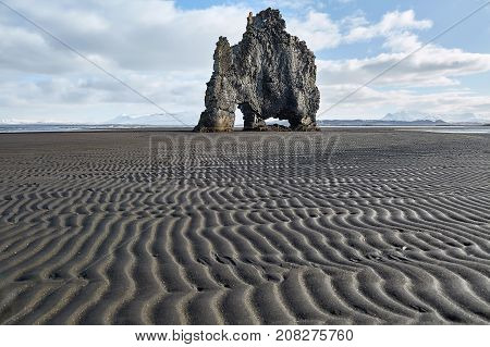 Abstract rock on the dark volcanic sand on the background of the blue sky with clouds. It is Hvitserkur during low tide in Iceland. Horizontal.