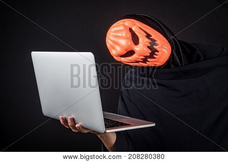 Anonymous male hacker with pumpkin halloween mask carrying laptop computer. Internet security and cyber attack concepts.