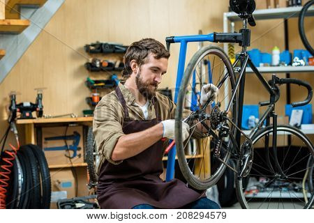 Young master in working gloves and apron repairing wheel of bicycle in workshop
