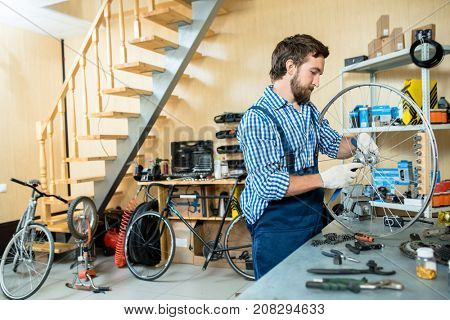 Young man in uniform and gloves repairing wheel of bicycle in workshop