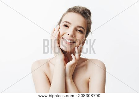 Cheerful young good-lookig student girl with dark long hair in bun hairstyle and naked body smiling with teeth, touching face with hands, looking in camera with sutisfied and relaxed face expression. Beauty and health.