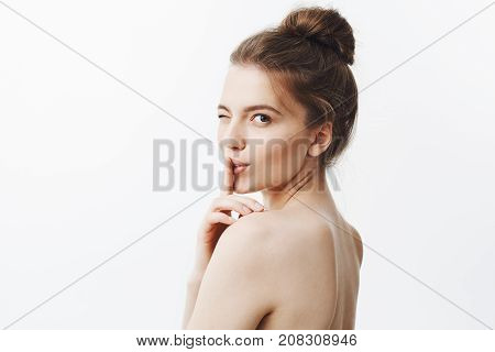Close up portrait of charming beautiful young european woman with dark long hair in bun hairstyle and naked skin, winking, touching lips with index finger in hush gesture, looking in camera with flirty and sexy face expression