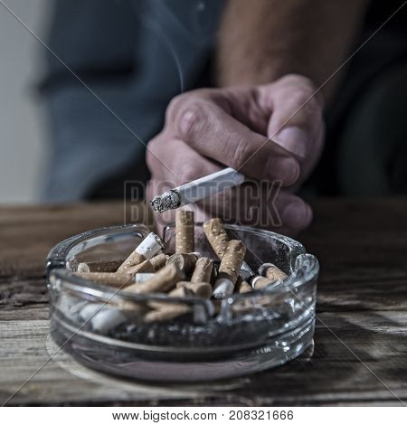 hand of man smoking cigarette with ashtray full of cig butt end and ash in disgusting unhealthy addiction and bad habit calling for quitting tobacco addiction and keep healthy lifestyle