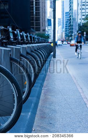 person riding a bike on a busy city street with cars and skyscrapers in the background and a row of wheel and bike at a bike rental station