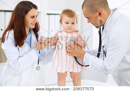 Happy cute baby at health exam at doctor's office. Toddler girl is standing while have been keeping by two doctors.