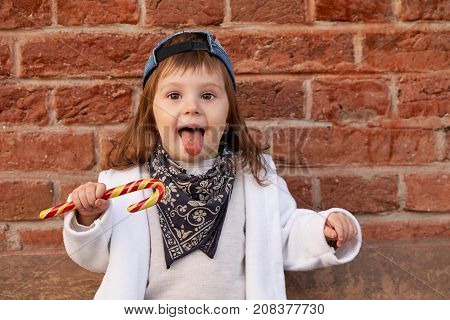 Cute beautiful child eats candy.little girl in denim hat shows tongue on the background of brick wall