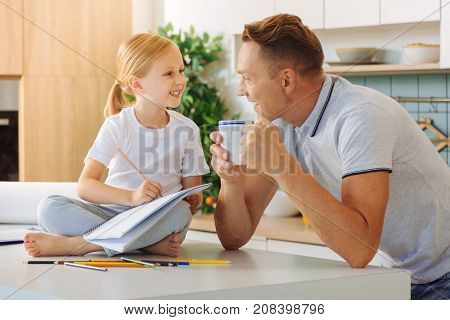 Interaction with daughter. Nice pleasant delighted man holding a cup of tea and smiling while talking to his daughter
