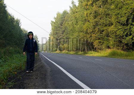 A boy in a dark hooded sweatshirt walks along the roadside in a beautiful forest