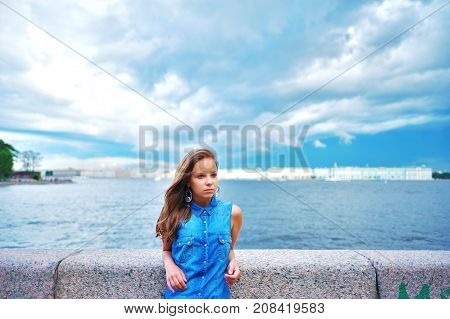 Portrait of young nice woman in a denim shirt with long luxurious hair, is on the St. Petersburg waterfront, leaning on the stone rail on blurred background of a beautiful sky, river and urban architecture.
