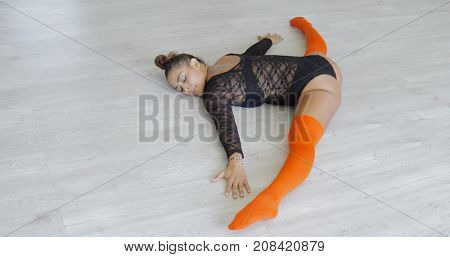 Content young dancer in orange stockings and black bodywear doing splits while warming up before training and lying on studio floor with eyes closed.