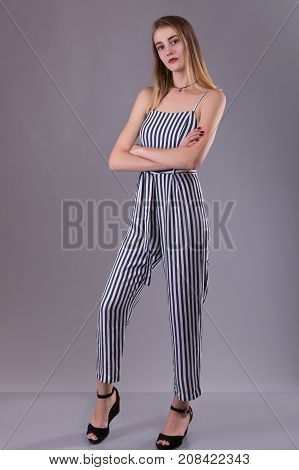 Confident attractive slender young woman wearing striped overall standing with folded arms looking thoughtfully at the camera over gray studio background with copy space