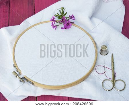 white fabric for embroidery in wooden round embroidery frame near needle scissors and thimble
