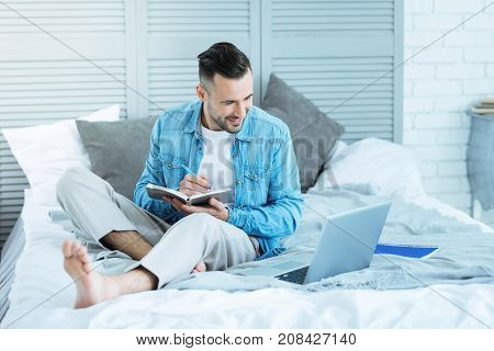 No time for worries. Cheerful millennial guy looking at a screen of his laptop with a smile on his face while sitting in bed and noting something down.