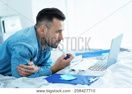The next big idea. Side view on a young entrepreneur looking at a screen of his laptop while relaxing on his bed and thinking about a new business idea at home.