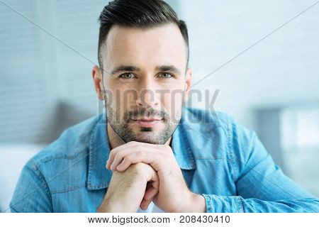 Successful and confident. Young hazel eyed gentleman resting his chin on his hands while spending his day indoors and looking into the camera.