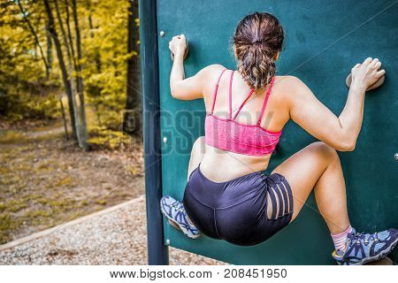 Back of young fit woman climbing rock bouldering wall in outdoor park in sports bra and shorts in autumn with foliage