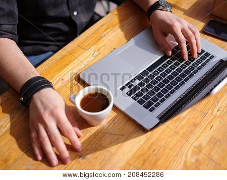 Fresh coffee for great ideas. A man working on a laptop and holding a cup of coffee while sitting at his workplace. Close-up.