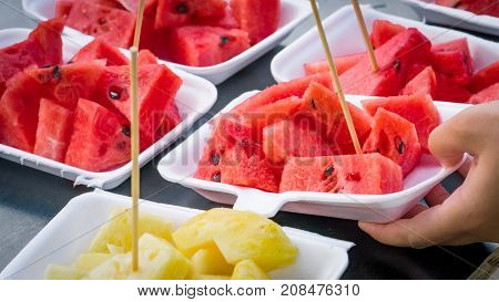 Fresh watermelon and pineapple slice in white foam plate container on black wooden table. Serving by one hand.