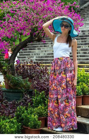 A girl in azure hat with fields a white tshirt and a long pink skirt against the background of a flower tree. A woman in Sanya, Hainan, China.