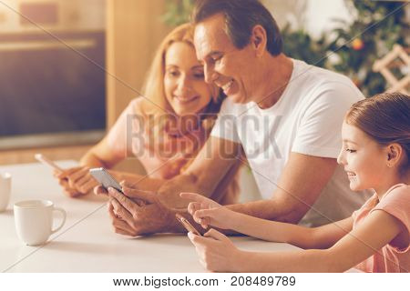 Harmonious relations. Selective focus on a beaming little girl sitting next to her father and grinning broadly while all using their smartphones and having a pleasant family conversation.