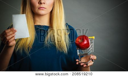 Worried woman holding shopping basket with fruits looking at bill receipt being scared of huge prices