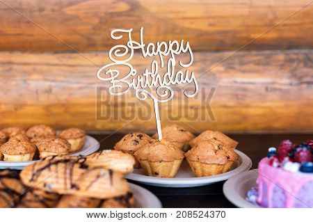 Birthday cake and muffins with wooden greeting sign on rustic background. Wooden sing with letters Happy Birthday and holiday sweets