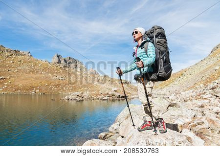 Hiker girl in sunglasses down jacket and hat with a backpack and mountain equipment with tracking sticks in her hands looks at the beautiful view of a high mountain lake in an archipelago in the northern Caucasus. Karachay-Cherkess Republic. 