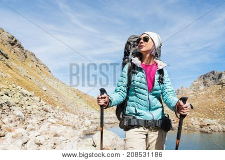 Hiker girl in sunglasses down jacket and hat with a backpack and mountain equipment with tracking sticks in her hands looks at the beautiful view of a high mountain lake in an archipelago in the northern Caucasus. Karachay-Cherkess Republic. 