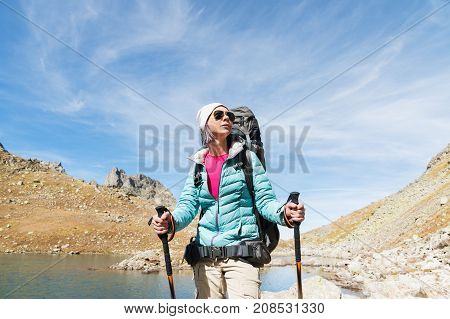 Hiker girl in sunglasses down jacket and hat with a backpack and mountain equipment with tracking sticks in her hands looks at the beautiful view of a high mountain lake in an archipelago in the northern Caucasus. Karachay-Cherkess Republic. 