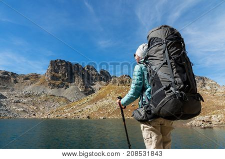 Hiker girl in sunglasses down jacket and hat with a backpack and mountain equipment with tracking sticks in her hands looks at the beautiful view of a high mountain lake in an archipelago in the northern Caucasus. Karachay-Cherkess Republic. The concept o