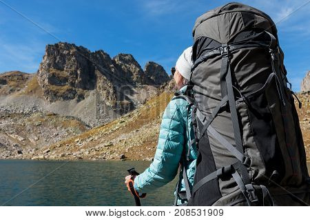Hiker girl in sunglasses down jacket and hat with a backpack and mountain equipment with tracking sticks in her hands looks at the beautiful view of a high mountain lake in an archipelago in the northern Caucasus. Karachay-Cherkess Republic. The concept o