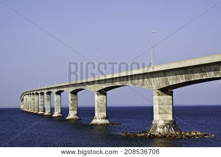 Long landscape view of the Confederation Bridge winding across to Prince Edward Island with seagulls on large rocks at the bridge's base on a sunny afternoon in August.