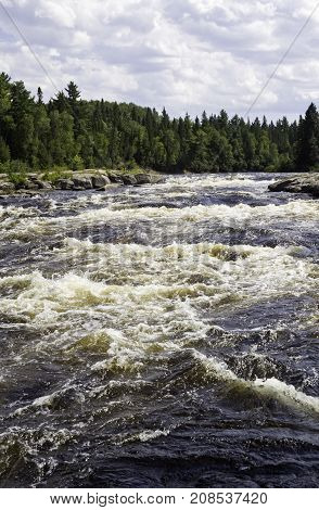 Vertical of rapids running between a rocky shoreline and surrounded by trees and greenery at Pabineau Falls near Bathurst, New Brunswick on a bright sunny day with blue skies and clouds in August.