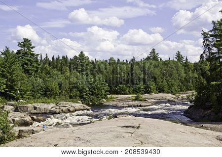 Wide view of a some small rapids off a rock shoreline surrounded by trees and greenery at Pabineau Falls near Bathurst, New Brunswick on a bright sunny day with blue skies and clouds in August.