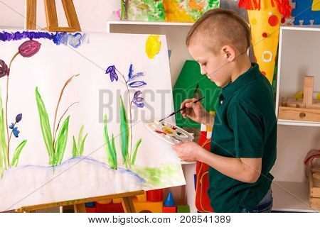 Child painting on easel. Kid boy learn paint by brush in class school. Kindergarten interior on background. Boy is getting ready to become an artist. He draw alone.