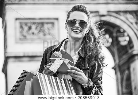 Stylish Christmas in Paris. Portrait of happy elegant woman in trench coat with shopping bags and Christmas present near Arc de Triomphe in Paris France