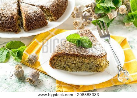 Delicious homemade poppy cake. Baking in multicooker. Whole pie and piece on yellow napkin mint and dry poppy capsule with seeds on table