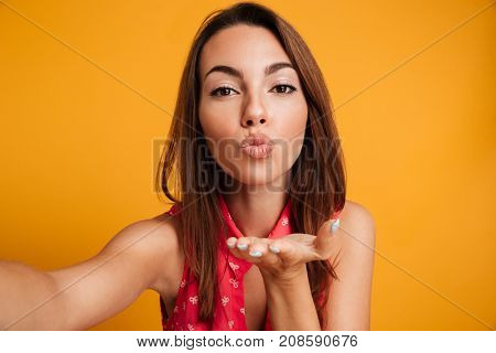 Pretty brunette woman in dress making selfie and sends air kiss at the camera over yellow background