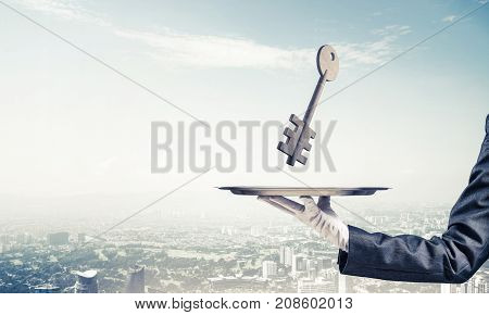 Closeup of waiter's hand in white glove presenting stone key symbol on metal tray with cityscape view on background. 3D rendering.