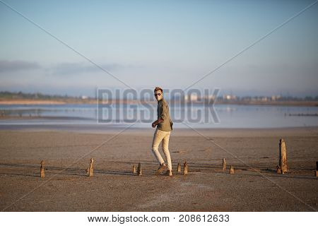 A young guy in white jeans, a shirt and glasses, on a sandy beach by the lake. Against the background of a blurry city. In full growth.