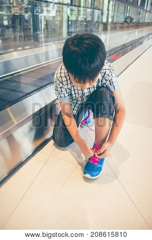 Asian Child Sitting And Tying To Tie Shoelaces Near Walkway In Modern Airport.