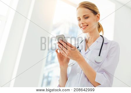 Taking a break. Low angle shot of a friendly looking female physician with a stethoscope smiling while focusing her attention on a screen of her smartphone and checking social medias.