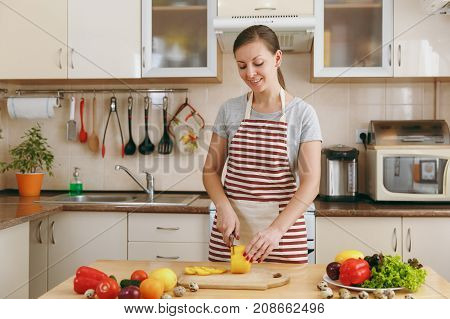 A Young Attractive Woman In An Apron Cuts Vegetables For Salad With A Knife In The Kitchen. Dieting
