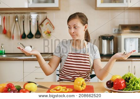 A Young Attractive Pensive Woman In An Apron Chooses Between Chicken And Quail Eggs In The Kitchen.