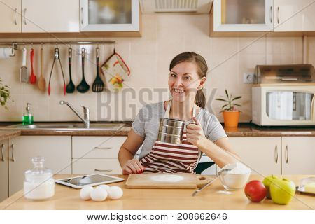 A Young Smiling Woman Sifts Flour With An Iron Sieve With Tablet On The Table In The Kitchen. Cookin