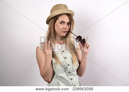 beautiful stylish young woman in grey denim dress a hat and sunglasses with a flirting expression on his face poses for a portrait on a white background