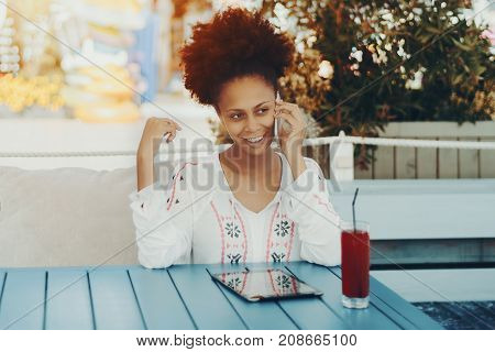 Portrait of skittish black girl sitting at the table of cafe outdoors and talking on the phone; smiling naughty young biracial woman in street bar with digital pad cell phone and glass of red juice
