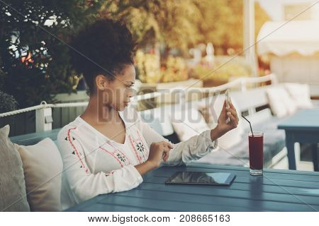 Young black female using front cam of her smartphone as mirror to check is everything okay with her face while sitting in street cafe with glass of red beverage and digital tablet and waiting friend