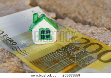 Small miniature white toy house with green roof stands on the banknote of euro lying on a beige stone.