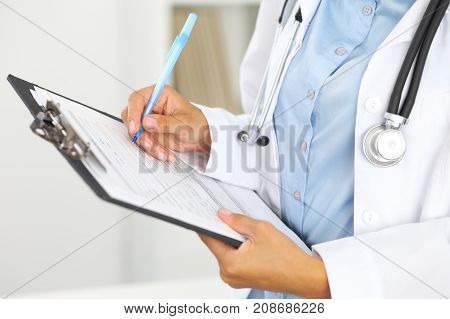 Close-up of a female doctor filling  up medical form at clipboard while standing straight in hospital.